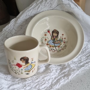 May include: A white ceramic mug and bowl with a floral design featuring a girl watering flowers. The mug has a handle and the bowl is shallow.