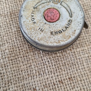Peut inclure: Un boîtier de mètre ruban circulaire en métal patiné avec une finition argentée et un petit bouton central rose. Le texte A1 est imprimé en haut, avec les mots MADE IN ENGLAND inscrits en arc de cercle sur la surface métallique.