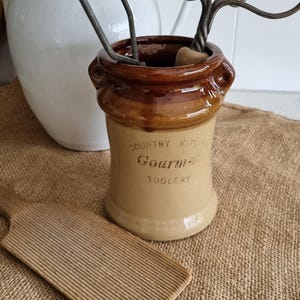 Puede incluir: Un recipiente de cerámica marrón y beige para utensilios de cocina con la inscripción "Country Kitchen Gourmet Toolery". Se ven una paleta de mantequilla de madera y batidores de metal.