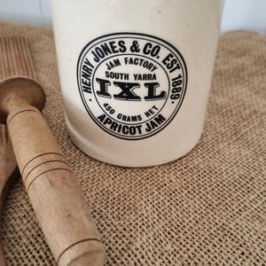 May include: A cream-coloured ceramic jar with a cork lid. The front features black text reading Henry Jones & Co. Est 1889, Jam Factory South Yarra, IXL, and 450 grams net Apricot Jam. The jar is cylindrical with a speckled finish and a flat base.