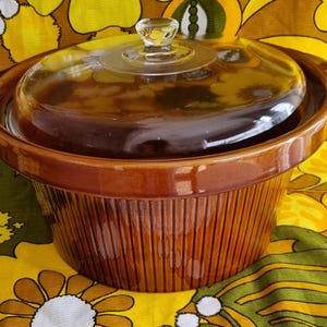 May include: A round, brown ceramic casserole dish with vertical ribbed detailing along the exterior sides. The dish includes a clear, circular glass lid featuring a central knob handle and a textured, concentric ring pattern on the top surface.
