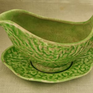 May include: A light green ceramic gravy boat with a textured, leaf-like surface pattern and a matching saucer. The vessel features a curved handle and a wide pouring spout, with subtle brown highlights accentuating the embossed details.