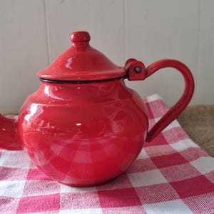 May include: A bright red teapot with a curved spout and a matching handle. The teapot has a lid and sits on a red and white checked cloth. The teapot is a classic kitchenware item.