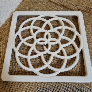 May include: White ceramic trivet with a geometric design. The design is a circle with a flower-like pattern inside.