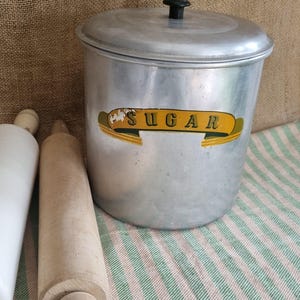 May include: A vintage aluminium sugar canister with a black knob on the lid and the word "SUGAR" on a yellow banner. Two wooden rolling pins are next to the canister. The items are on a striped green and white cloth.