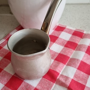 May include: A small, silver-colored metal pot with a long handle, resting on a red and white checkered cloth. A white ceramic pitcher is in the background. The pot appears to be for heating liquids.