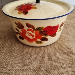 May include: A cream-coloured enamel bowl with a matching lid featuring a black knob. The bowl and lid are decorated with painted red and orange floral patterns with green leaves. The rim of the bowl is accented with a thin dark blue line.