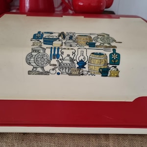 May include: A rectangular bread box with a red base and a cream-colored roll-top lid. The lid features a central graphic print in blue, yellow, and black, depicting various kitchen items including a coffee grinder, a barrel, and a kettle.