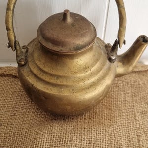May include: A brass teapot with a rounded body, a short spout, and a tall, arched handle. The teapot features a removable lid with a small central knob. The metal surface shows a weathered, aged patina with visible signs of wear and oxidation.