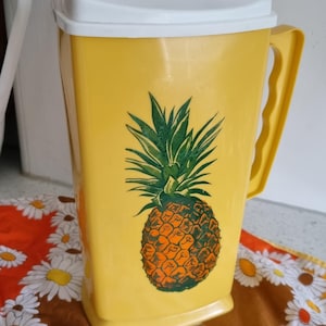 May include: Yellow plastic jug with a white lid and handle. The jug features a large pineapple graphic in shades of green and orange. The jug is sitting on a floral patterned cloth.
