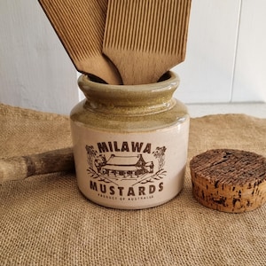 May include: A ceramic jar with a tan base and a green rim, featuring the brown text MILAWA MUSTARDS and a house illustration. Two wooden butter paddles are inside the jar, and a round cork stopper sits to the right of the jar on the surface.
