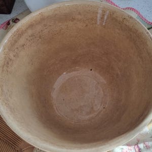 May include: A large, round ceramic mixing bowl with a smooth, light tan interior featuring subtle brown speckles and a glossy finish. The bowl has a wide, open top and deep, curved sides, showcasing a consistent, neutral-toned earthenware construction.