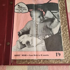 May include: A vintage Patons Knitting Book No. 533, showing a baby in a knitted cardigan. The cover is pink with black and white illustrations and text. The book is open in a red binder, showcasing a classic baby wear pattern.