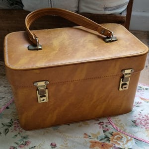 May include: A vintage tan suitcase with a handle and two metal clasps. The rectangular case has a slightly worn appearance, suggesting age and use. The handle is a matching tan colour, and the clasps are gold-toned.