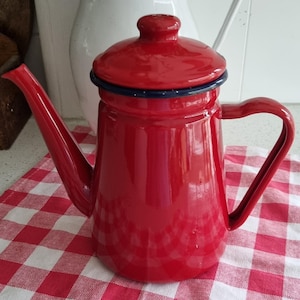 Puede incluir: Una cafetera de esmalte rojo brillante con tapa a juego y asa curvada. La cafetera tiene un pico largo y un borde azul oscuro. Se encuentra sobre un mantel a cuadros rojos y blancos, evocando un ambiente vintage.