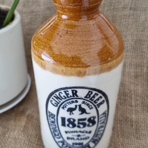 May include: A ceramic ginger beer bottle with a two-tone design featuring a brown glazed neck and a cream-coloured body. The front displays a black circular stamp with the text Ginger Beer, Potters Since 1858, Bendigo Pottery Australia, Pinnacle Brand.