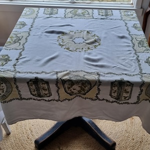 May include: A square white tablecloth featuring a repeating border pattern of green and beige illustrations of teapots, tea canisters, and plates. The center of the fabric displays a circular floral wreath design in muted green and beige tones.