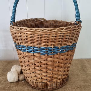 May include: A round woven wicker basket featuring a natural light brown finish with a metallic blue accent band woven into the upper section and a matching metallic blue wrapped handle. Three small white yarn balls sit near the base.