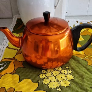 May include: A polished, orange-toned teapot with a black handle and lid. The teapot is on a patterned tablecloth with yellow and green floral designs. A white jug is in the background.