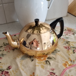 May include: A gold teapot with a black handle and a shiny, reflective surface. The teapot is sitting on a floral patterned tablecloth.