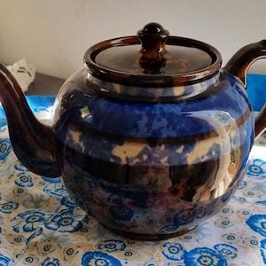 May include: A round, ceramic teapot with a dark brown lid, handle, and spout. The body of the teapot is a deep blue with brown and cream-coloured horizontal stripes. The teapot sits on a floral patterned fabric.
