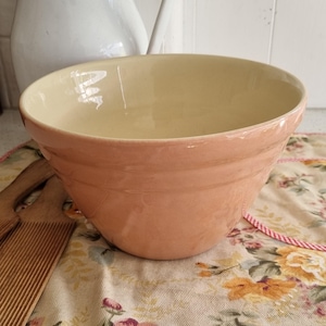 May include: A large, peach-colored ceramic mixing bowl with a cream-colored interior. The bowl has a rounded shape with a slightly flared rim and a ridged exterior. The bowl sits on a floral patterned cloth.