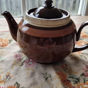 May include: A round ceramic teapot featuring a glossy finish with horizontal bands in shades of dark brown, medium brown, and cream. The teapot includes a matching lid with a small knob, a curved spout, and a sturdy handle on the side.