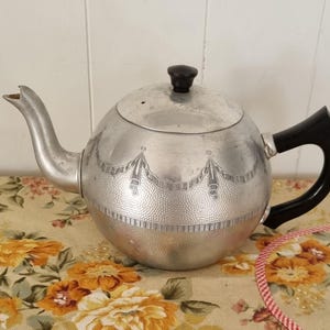 May include: A round, silver-coloured metal teapot featuring a textured lower body and a smooth upper section with an engraved swag and ribbon pattern. It includes a curved spout, a black handle, and a matching black knob on the removable lid.