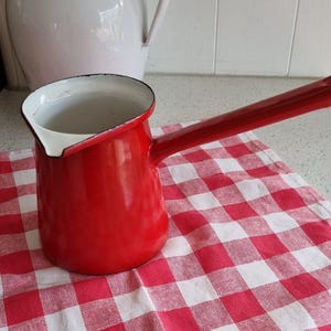 May include: A red enamel coffee pot with a long handle and a white interior. The pot sits on a red and white checked tablecloth. A white jug is in the background.