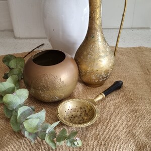 May include: A collection of antique brass items displayed on a hessian surface. Features a tall, ornate jug, a spherical bowl, and a tea strainer with a black handle. Eucalyptus branches provide a natural accent. The items have etched floral patterns.