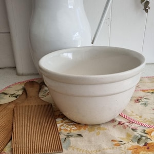 Op de afbeelding: Een crèmekleurige keramische mengkom met een ronde vorm en twee horizontale banden. Een witte kan en twee houten boterplanken zijn ook zichtbaar. De items staan op een bloemmotiefstof.
