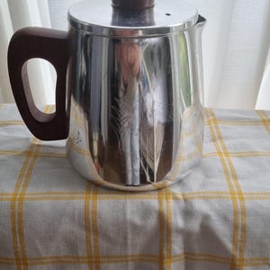 May include: A silver coffee pot with a wooden handle and lid knob. The pot is sitting on a white and yellow checked tablecloth.