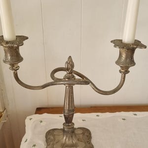 May include: A silver-toned metal candelabra featuring two arms with candle holders, each containing a long white taper candle. The central stem is fluted, rising from a square, ornate base with embossed floral details and a weathered finish.