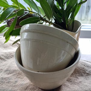 May include: Two stacked cream-colored ceramic mixing bowls with a smooth, glossy finish and a subtle horizontal ridge detail around the upper exterior. The bowls feature a simple, rounded shape and a neutral off-white tone throughout the surface.