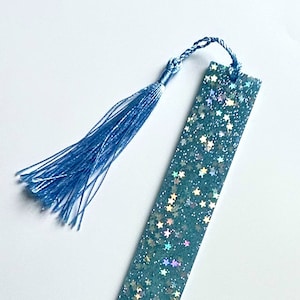 May include: A teal resin bookmark with embedded star-shaped glitter. The bookmark has a matching blue tassel attached at the top. The rectangular bookmark is approximately 6 inches long.