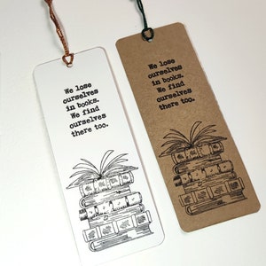 Puede incluir: Dos marcapáginas con una pila de libros y el texto "We lose ourselves in books. We find ourselves there too." Un marcapáginas es blanco con texto negro y una ilustración en blanco y negro. El otro marcapáginas es marrón con texto negro y una ilustración en blanco y negro.