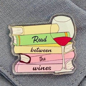 Op de afbeelding: Een transparante acryl boekenlegger met een stapel kleurrijke boeken en een glas rode wijn. De boekenlegger heeft de tekst "Read between the wines" erop.