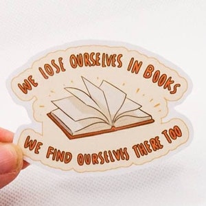 Puede incluir: Una pegatina blanca con una ilustración de un libro abierto y el texto "We lose ourselves in books. We find ourselves there too."
