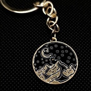Puede incluir: Llavero dorado con un dije redondo de esmalte negro que presenta una imagen estilizada de una cordillera, una luna creciente y estrellas.