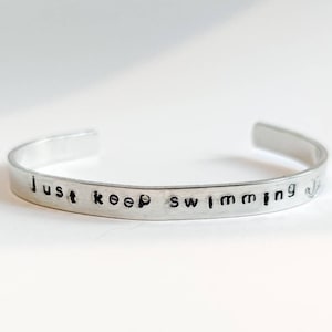 Könnte beinhalten: Ein silbernes Armband mit dem geprägten Satz "Just keep swimming". Das offene Armband besteht aus einem glänzenden, metallischen Material und wird auf einer weißen Oberfläche präsentiert. Das Armband ist ein schlichtes, aber elegantes Schmuckstück.