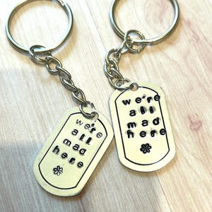 Puede incluir: Dos llaveros plateados con un dije de placa de identificación plateada. Cada placa tiene el texto "we're all mad here" en letras negras.