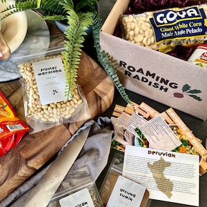 May include: A brown cardboard box with the words "Roaming Radish" printed on it. The box contains a variety of Peruvian ingredients, including a bag of white beans, a bag of corn, a jar of green sauce, and a bag of ground cumin. The box also includes recipe cards for Peruvian dishes.