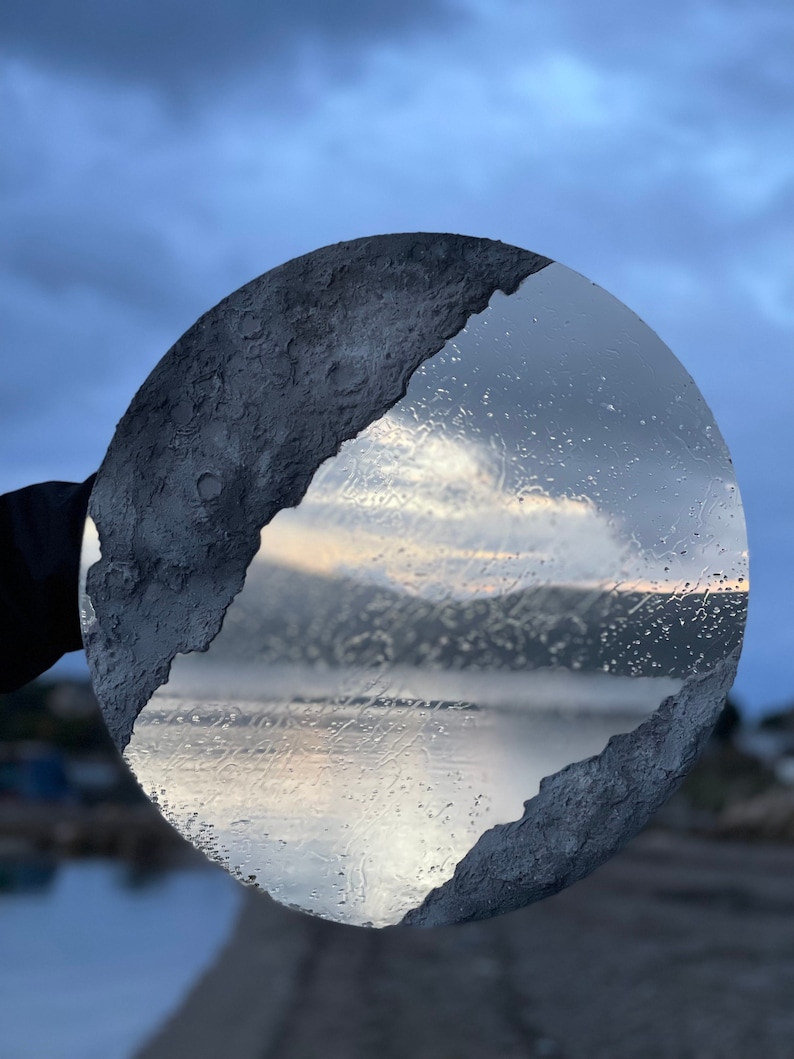 May include: A round, gray, textured, decorative wall hanging with a cracked, mirrored surface reflecting a cloudy sky and a body of water.