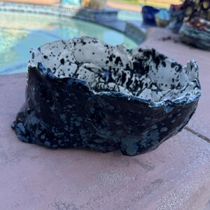 May include: A handmade ceramic bowl with a dark blue exterior and a speckled white and black interior. The bowl has a rough, textured surface and an irregular, organic shape. The bowl is sitting on a pink surface.