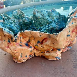 May include: A large, hand-formed ceramic bowl with an irregular, ruffled rim. The exterior features a light peach base with scattered blue and red speckles, while the interior is finished in a deep, mottled teal glaze with similar color accents.