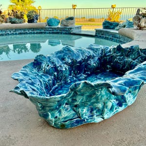 May include: A large, oval-shaped ceramic bowl with a highly textured, undulating rim. The surface features a glossy, multi-tonal glaze in shades of deep ocean blue, turquoise, and teal, mimicking the appearance of crashing waves.