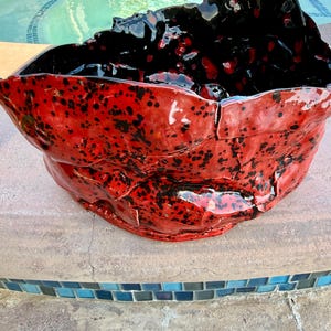 May include: A large, handcrafted ceramic bowl with a unique, organic shape. The bowl features a vibrant red exterior speckled with black spots, and a glossy black interior. The rim has a textured, uneven edge, adding to its artistic appeal.