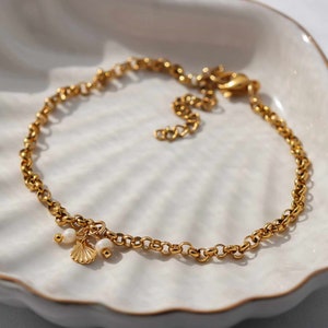 Könnte beinhalten: Ein goldenes Fußkettchen mit einem kleinen Muschel-Anhänger und zwei weißen Perlen.