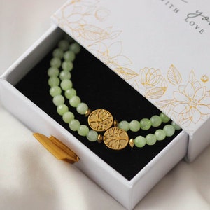 Puede incluir: Un brazalete de piedra de jade verde con dos dijes de árbol de la vida de oro en una caja de regalo blanca con un patrón floral y el texto "with love".