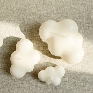 May include: Three white cloud-shaped candles of different sizes on a beige textured surface.